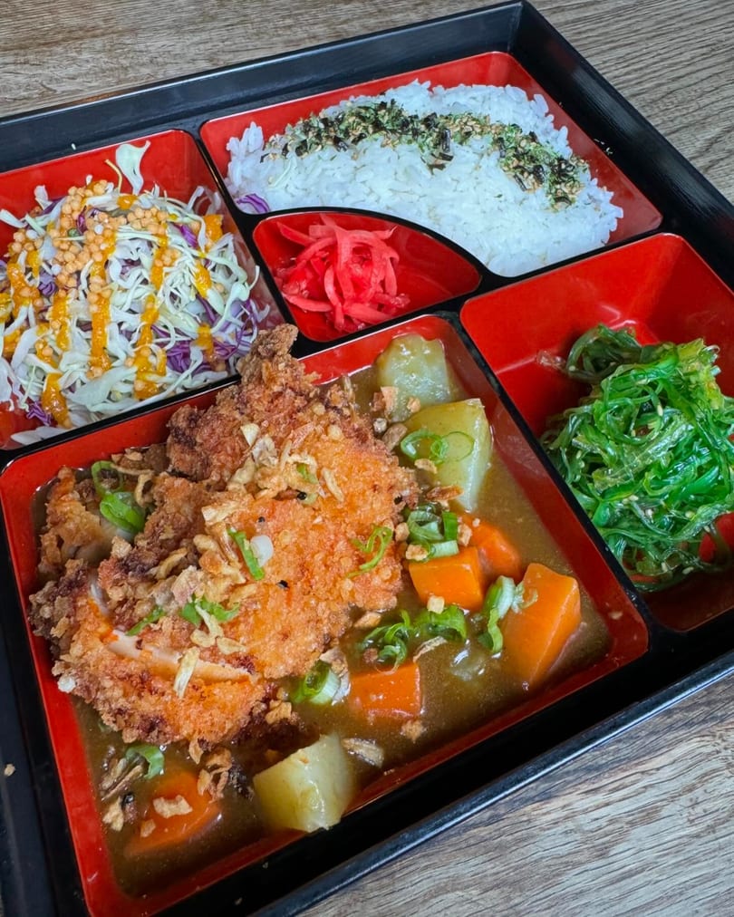 Japanese bento box with chicken katsu curry, rice, cabbage salad, seaweed salad, and pickled ginger.