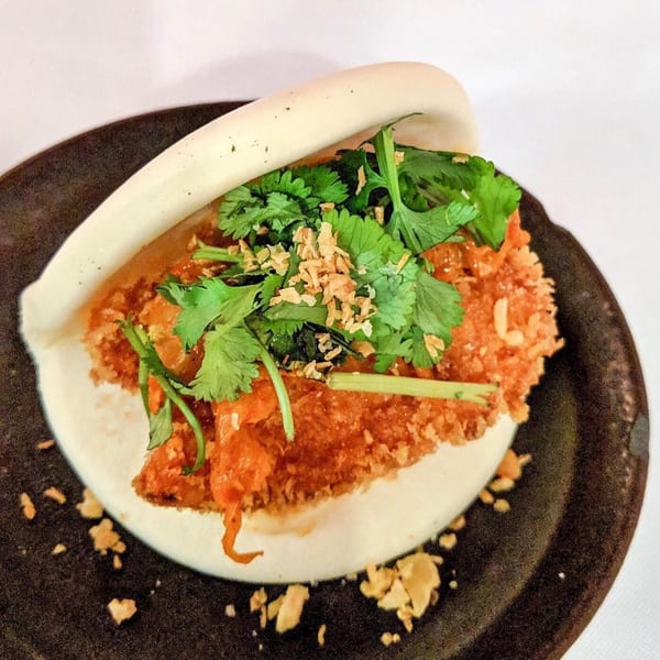 Crispy fried chicken bao bun with fresh cilantro and toasted garlic on a dark plate.