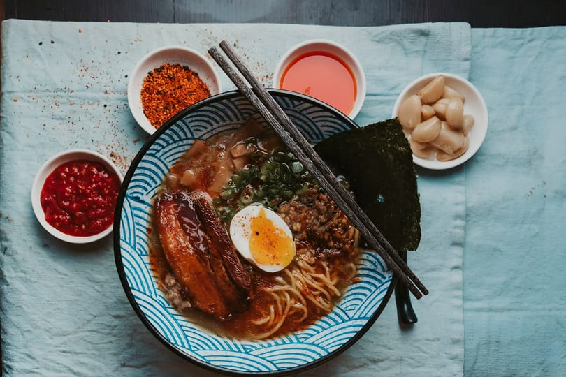 A bowl of ramen with pork belly, egg, noodles, and seaweed, surrounded by condiments.