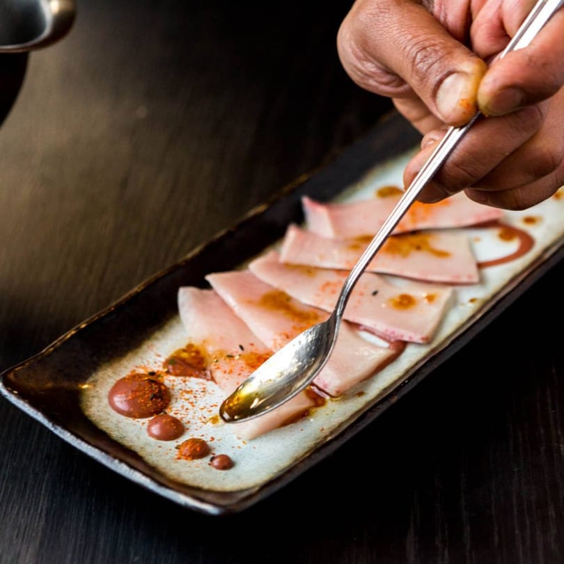 A chef's hand drizzles sauce onto thinly sliced fish on a rectangular plate.