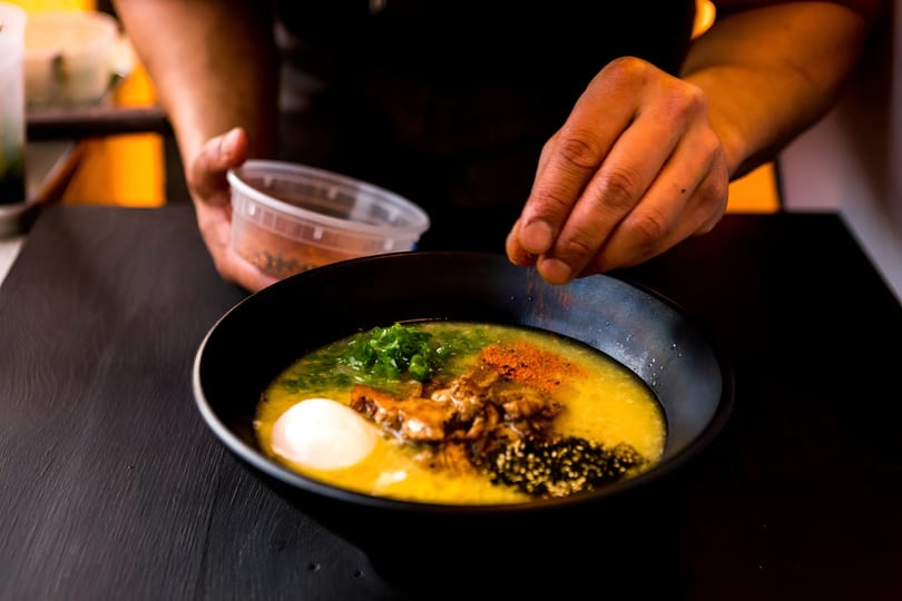 A chef's hands sprinkle spices over a vibrant bowl of ramen with egg and green onions.