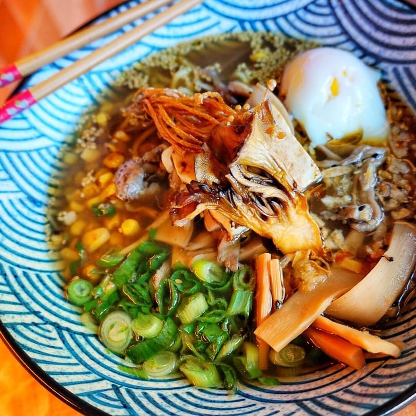 Close-up of ramen with mushrooms, scallions, corn, and a poached egg in a patterned bowl.