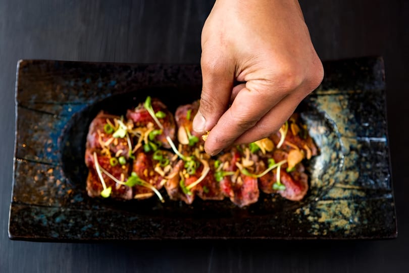 A hand sprinkles garnish over thinly sliced beef tataki on a dark rectangular plate.