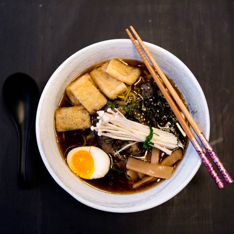 A bowl of ramen with tofu, mushrooms, egg, and chopsticks on a dark surface.